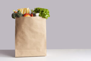 Various grocery items in paper bag on white table opposite gray wall. Bag of food with fresh vegetables, fruits, pasta and canned goods.