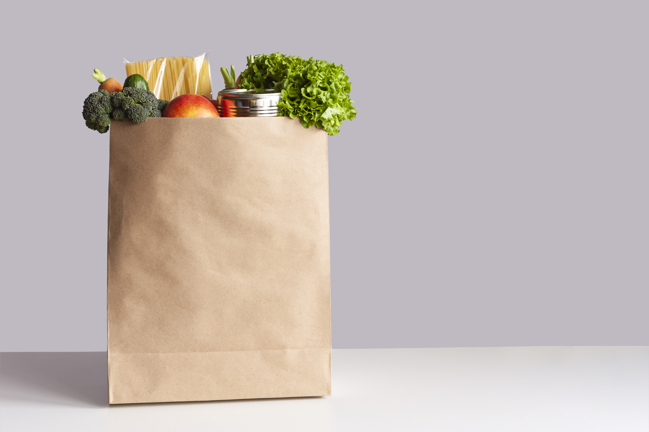 Various grocery items in paper bag on white table opposite gray wall. Bag of food with fresh vegetables, fruits, pasta and canned goods.