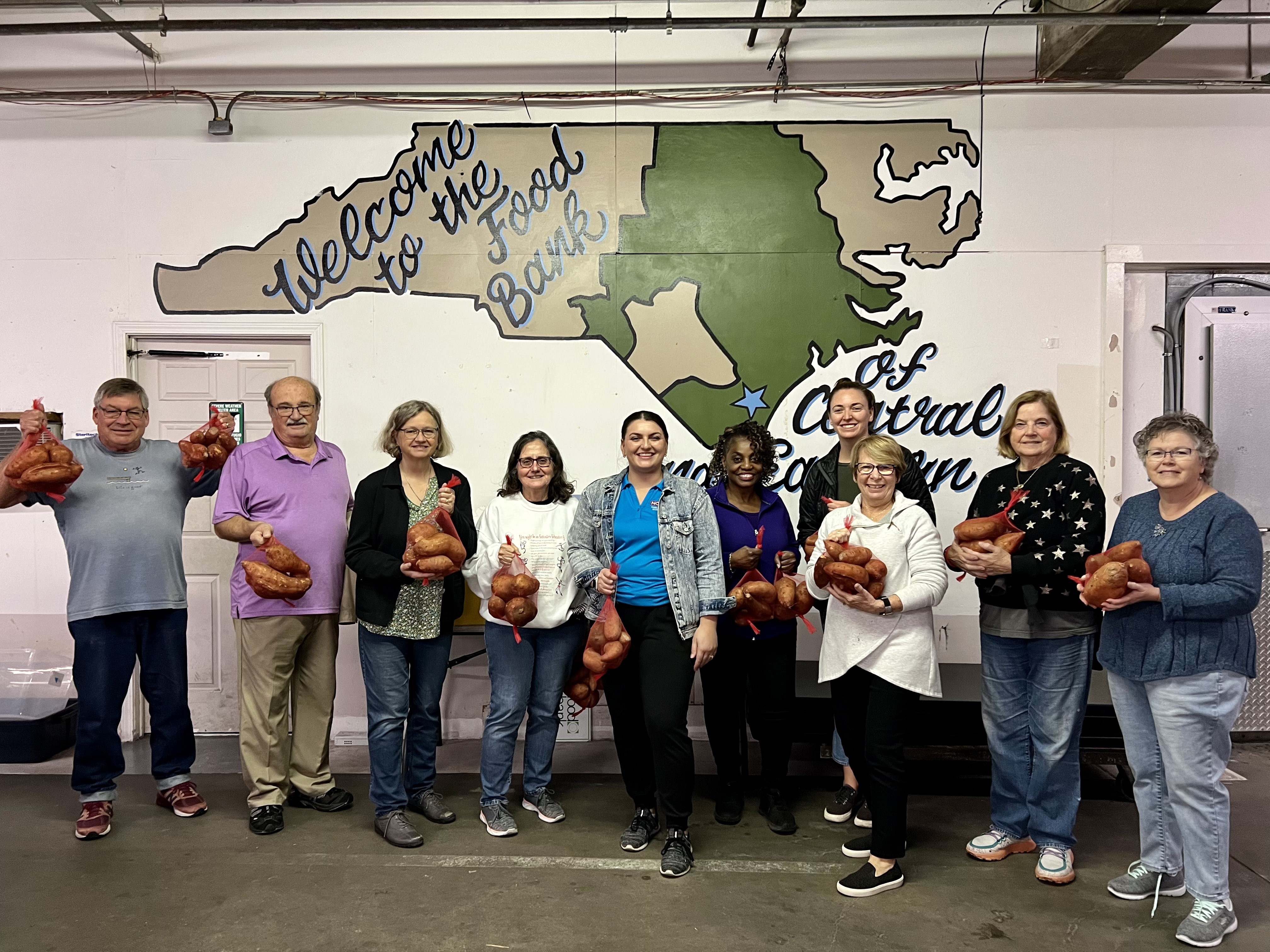 A group of people in front of a mural that reads "Welcome to the Food Bank of Central Carolina.