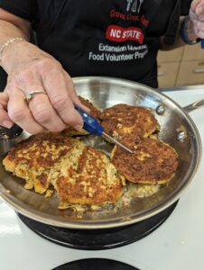 salmon burger patties being cooked in a skillet on a stove. Extension Master Food Volunteer is checking the temperature of the patties with a thermometer.