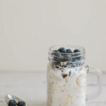 Vertical photo of overnight oats with coconut milk and blueberry served on wooden table with a metal spoon. Porridge in a jar. Breakfast meal