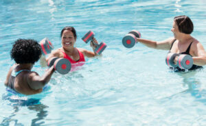 A multi-ethnic group of three senior women in their 60s doing water aerobics. The African-American woman and her two Hispanic friends are working out dumbbells, standing in shoulder-deep water.